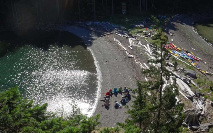 From an aerial point of view, a group of people sit in a circle on a beach. Kayaks and driftwood are also beached nearby.